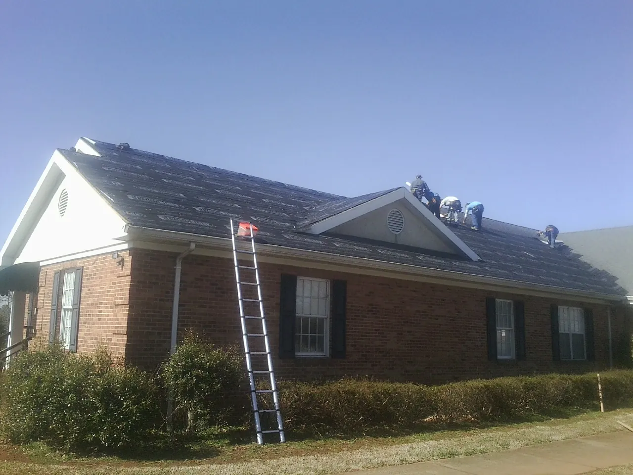 Crew tearing shingles off a brick ranch roof with a ladder set — Metal Roof Repair work by our crew in Statham