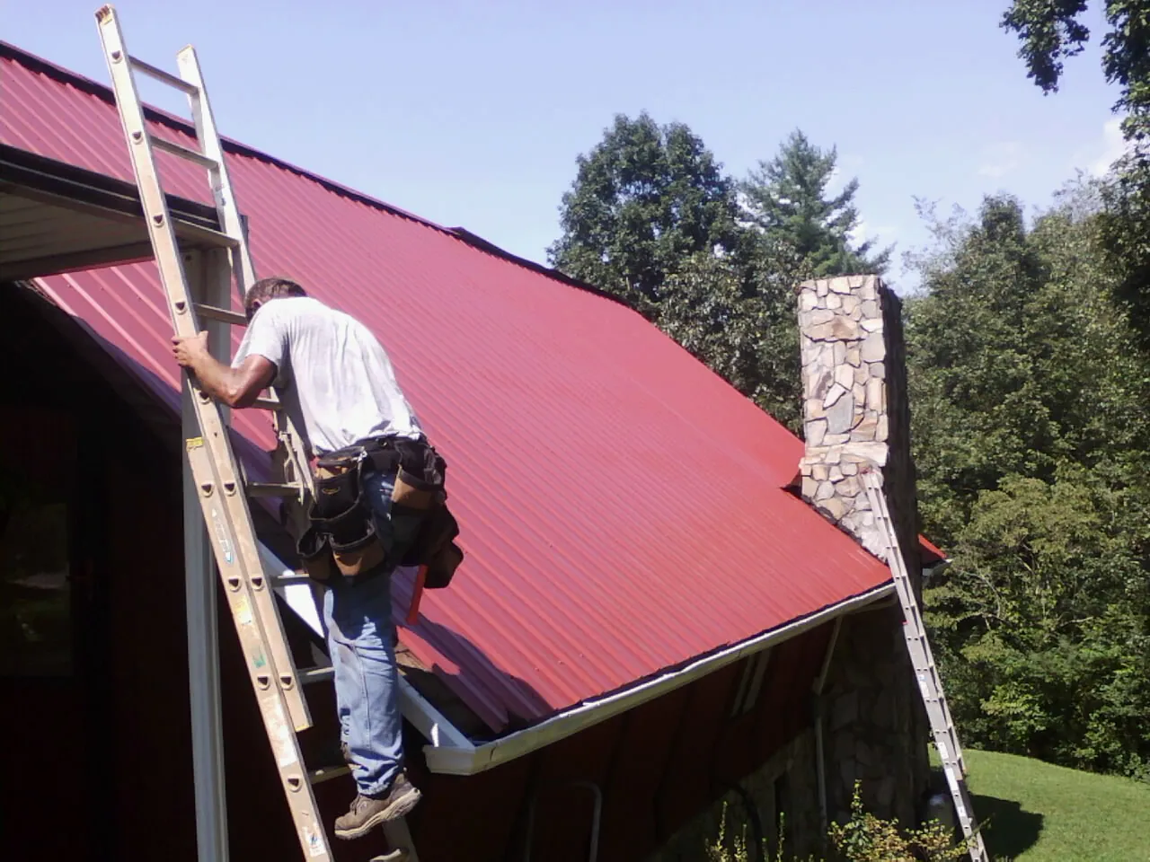 Roofer on a ladder beside a red metal roof and stone chimney — Soffit & Fascia Repair by Roofing Brothers in Statham